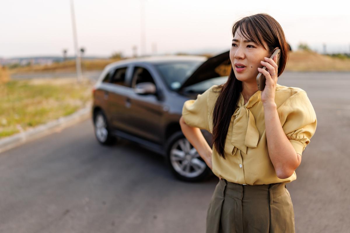 Woman stands by her broken-down car on the roadside with its hood open, making an urgent phone call.