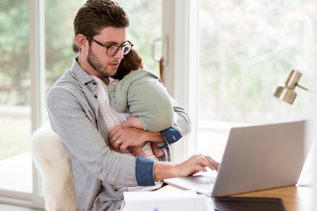 Man holding an infant while using a laptop at a desk at home.