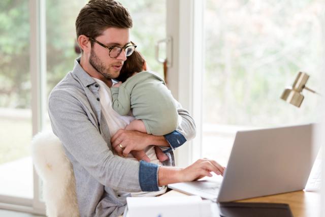 Man holding an infant while using a laptop at a desk at home.