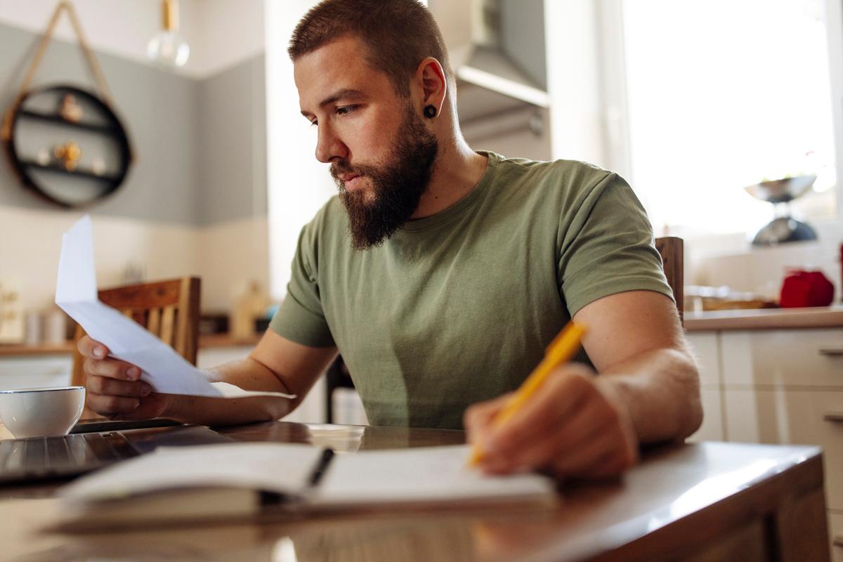 Man reviewing bills and finances in kitchen at home and taking notes with notebook and pen.