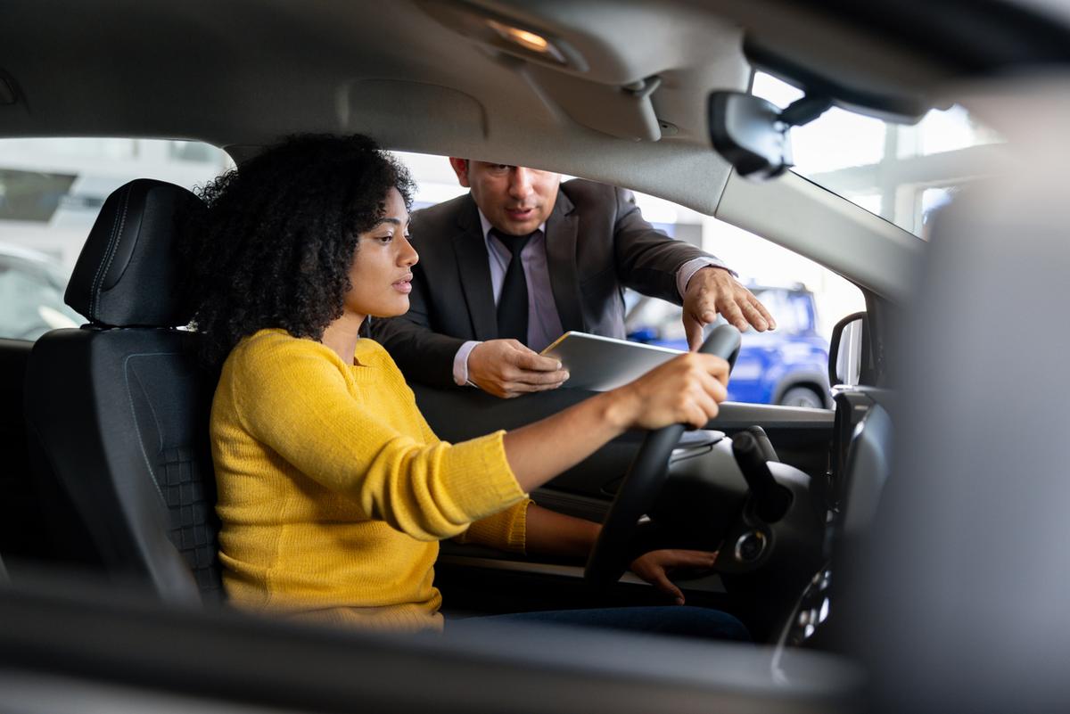 Woman sitting in driver's seat of new car talking to salesman at dealership.