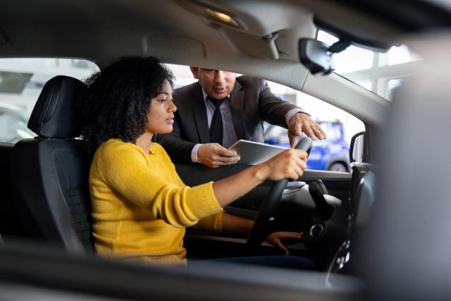 Woman sitting in driver's seat of new car talking to salesman at dealership.
