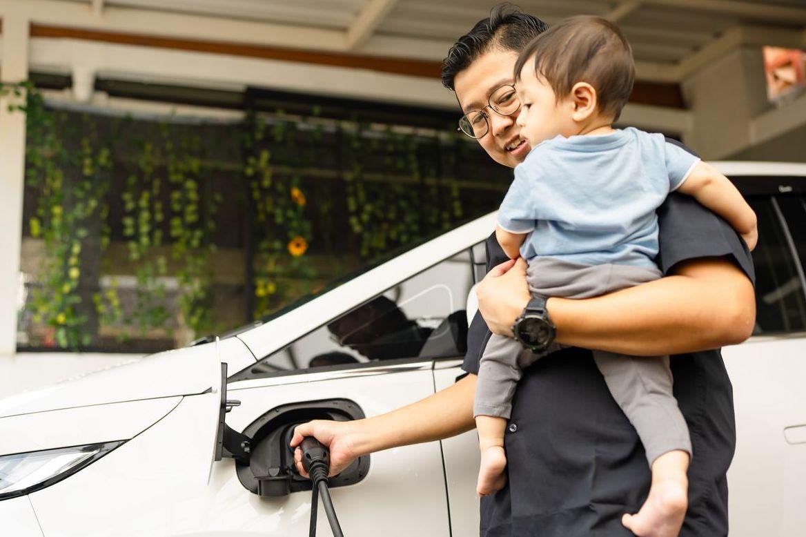 Father holds his young son while plugging an electric vehicle into the charging port at their home.