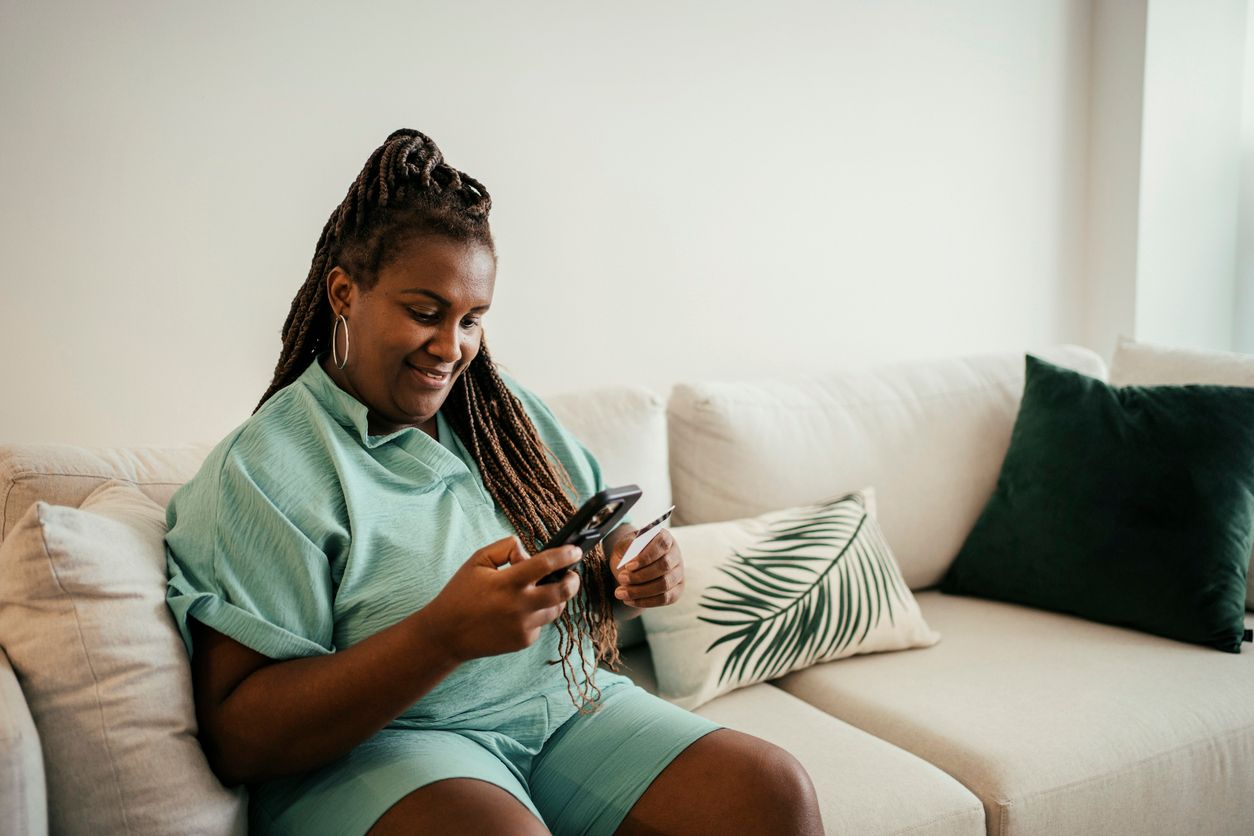 Woman at home on couch looking at phone and credit card.