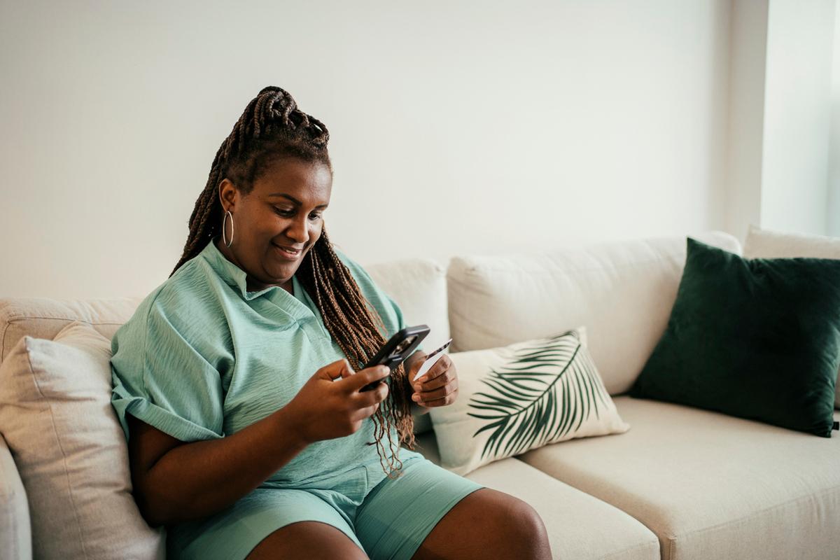 Woman at home on couch looking at phone and credit card.
