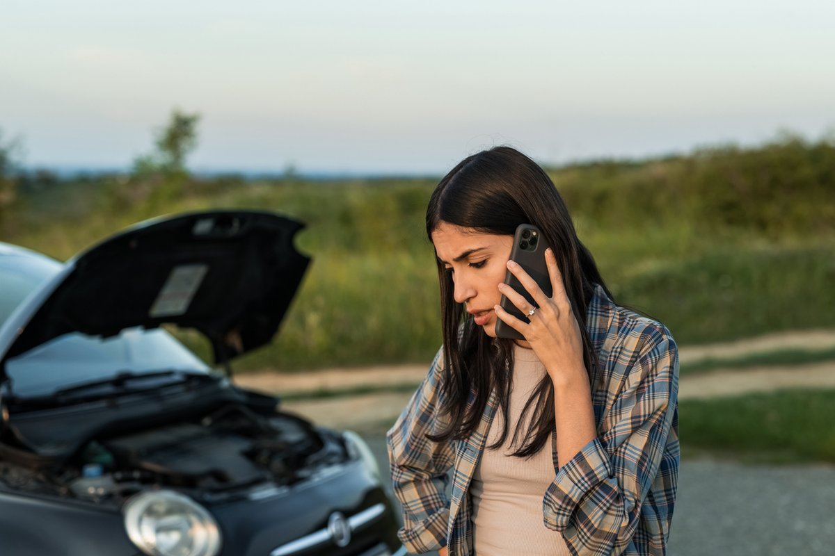Woman standing in front of a broken car and using phone to call for help.