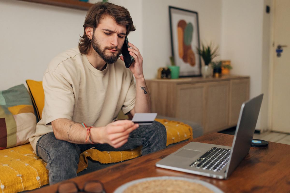 Young man with laptop at home, looking at credit card and calling his bank.