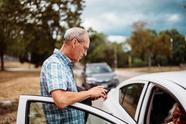 A senior man stands beside his car, using his phone while his wife is in the passenger seat.