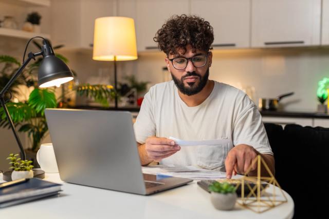 Man reviewing bills at kitchen table at home while using laptop.