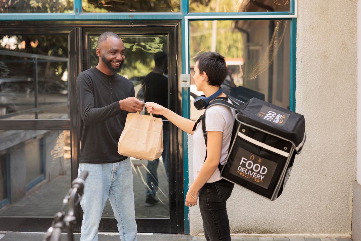 Food delivery service, young woman courier giving client lunch paper bag.