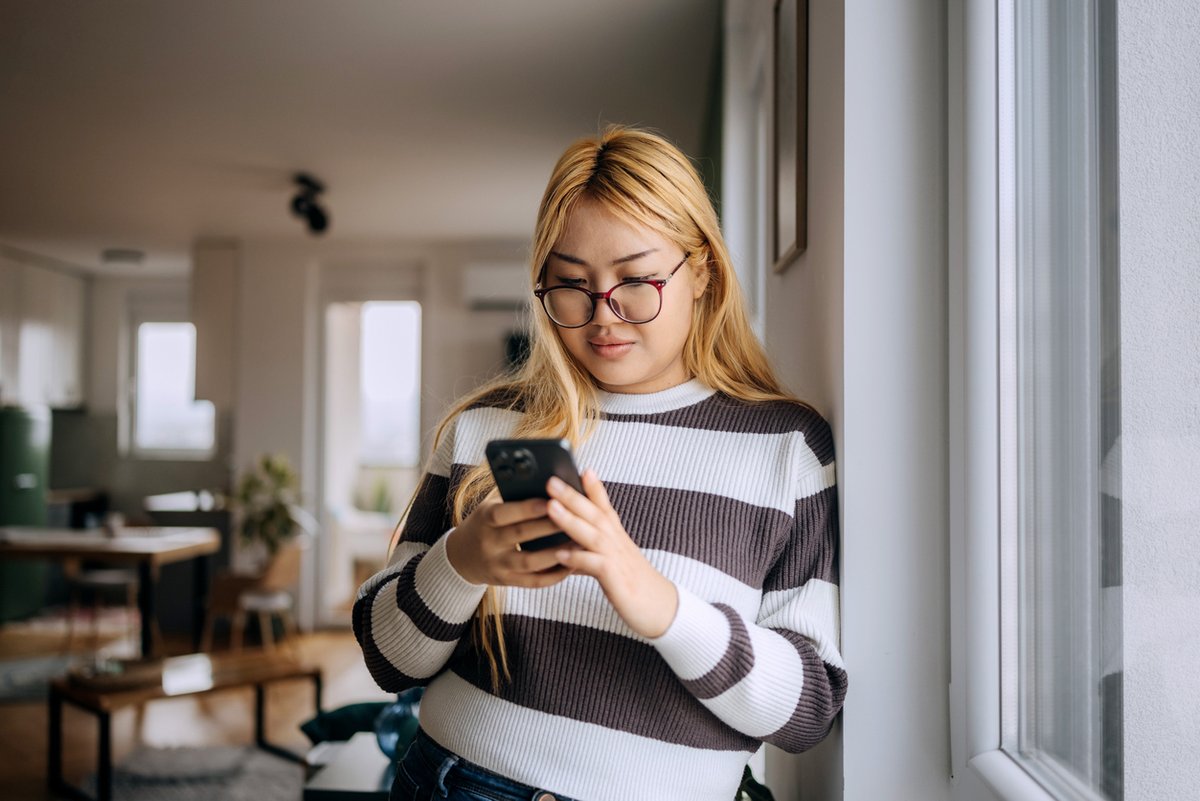 Young woman leaning against wall looking at phone at home.